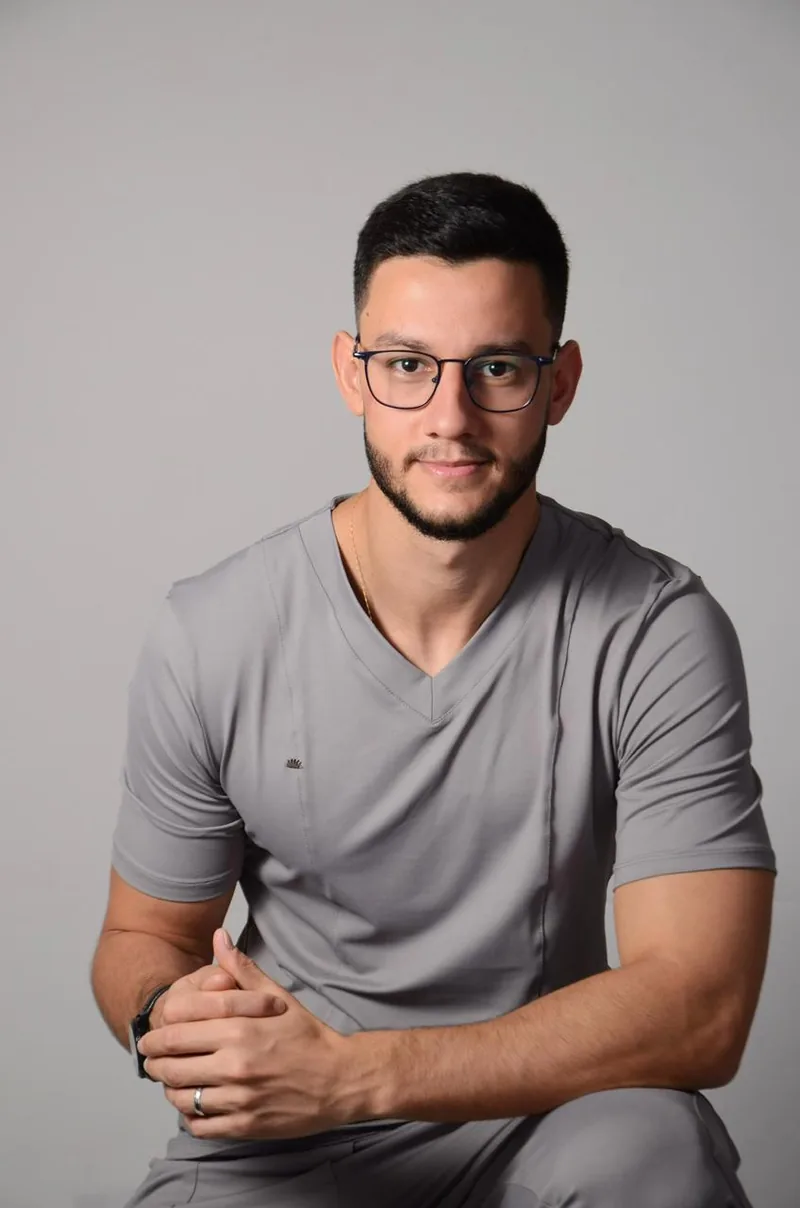 Dr. João Pedro Neves, dentista responsável pela Brasil Implantes, sentado, de uniforme clínico cinza e óculos